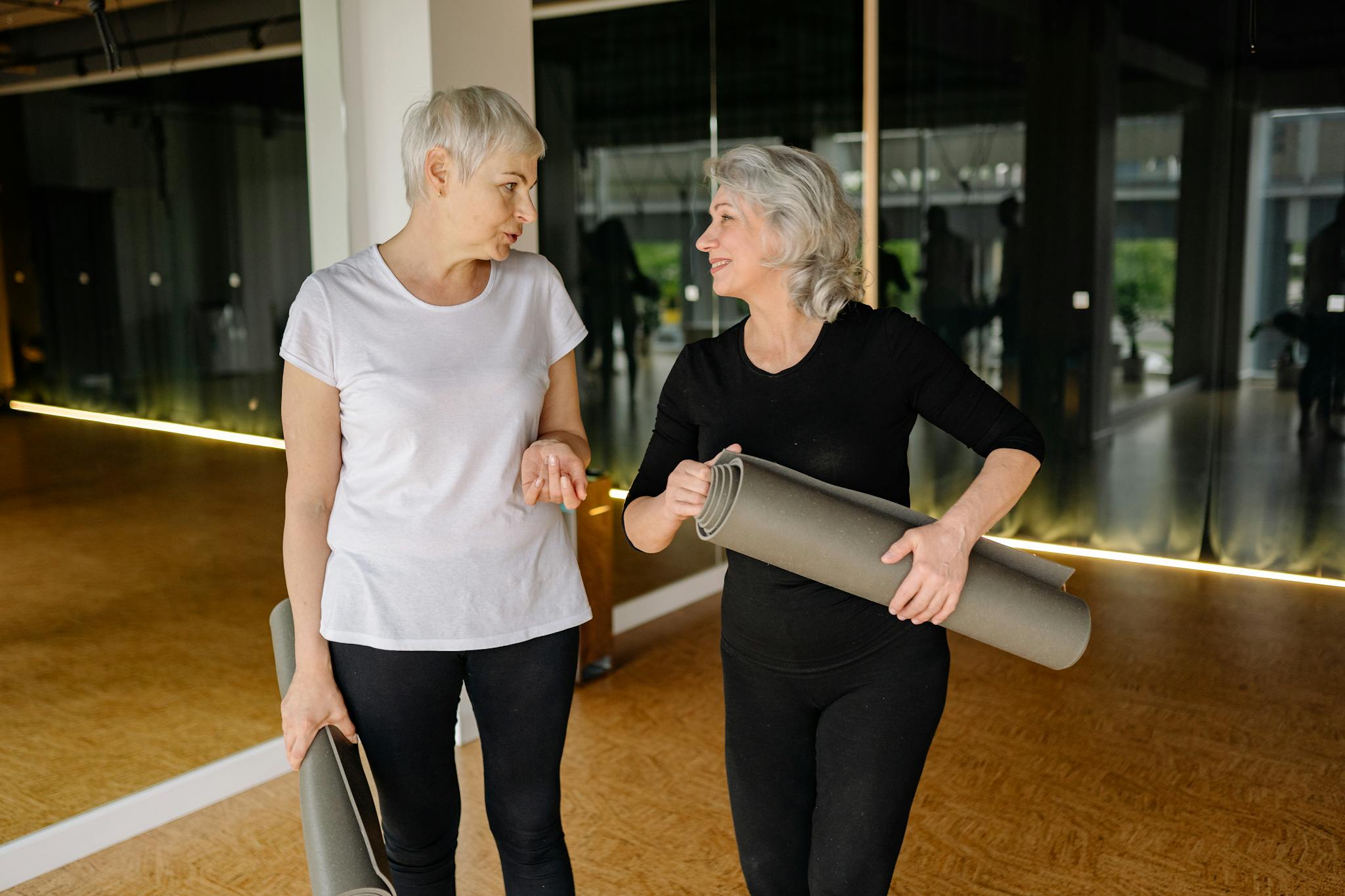 Two senior women engaging in conversation while holding yoga mats inside a fitness studio.